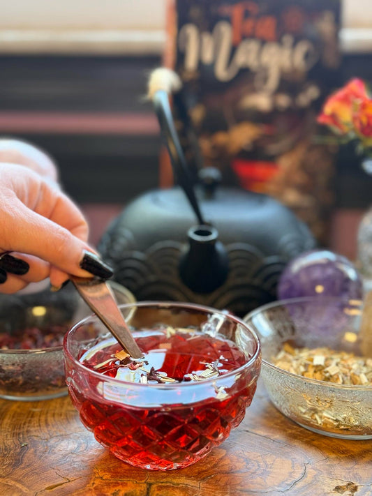 "Hand stirring a vibrant loose leaf tea in a crystal cup during a witchy tea ritual with herbs, crystals, and cast iron teapot for manifestation magic.