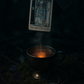 A mystical photo of Cream Earl Grey tea poured into a vintage silver cup, surrounded by moss, a runed silver charm, and a floating High Priestess tarot card—capturing the essence of Ancient Whispers tea blend inspired by deep intuition and hidden knowledge