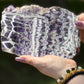 Polished chevron amethyst slab held outdoors, showcasing vivid purple and white zigzag patterns with quartz layering and raw edges.