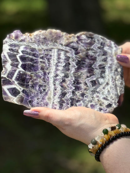 Polished chevron amethyst slab held outdoors, showcasing vivid purple and white zigzag patterns with quartz layering and raw edges.