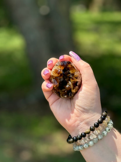 Cut base dark raw citrine crystal cluster displayed in hand with gemstone bracelets in natural sunlight