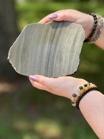 Full view of a Golden Obsidian slab with polished front and raw edges, showing the stone's chatoyant golden sheen against a natural outdoor backdrop.