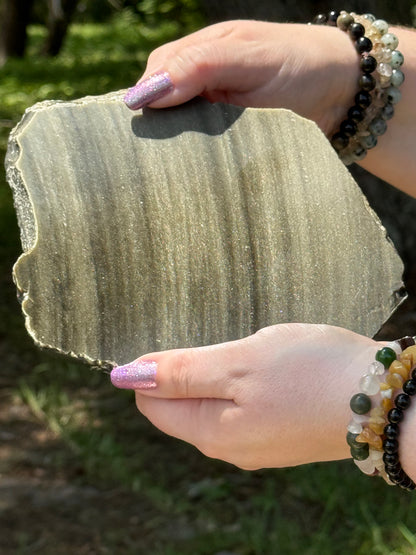 Close-up of a polished Golden Obsidian slab with visible golden shimmer and raw edges, displayed in natural sunlight.