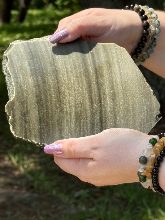 Close-up of a polished Golden Obsidian slab with visible golden shimmer and raw edges, displayed in natural sunlight.