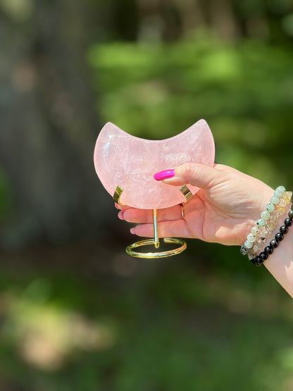 Hand holding a Rose Quartz crescent moon on a gold stand, symbolizing love and heart chakra healing in natural light