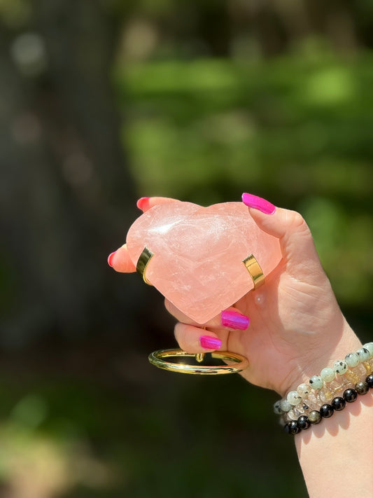 Rose Quartz heart-shaped crystal on gold stand held in hand outdoors, symbolizing love, healing, and Heart Chakra energy